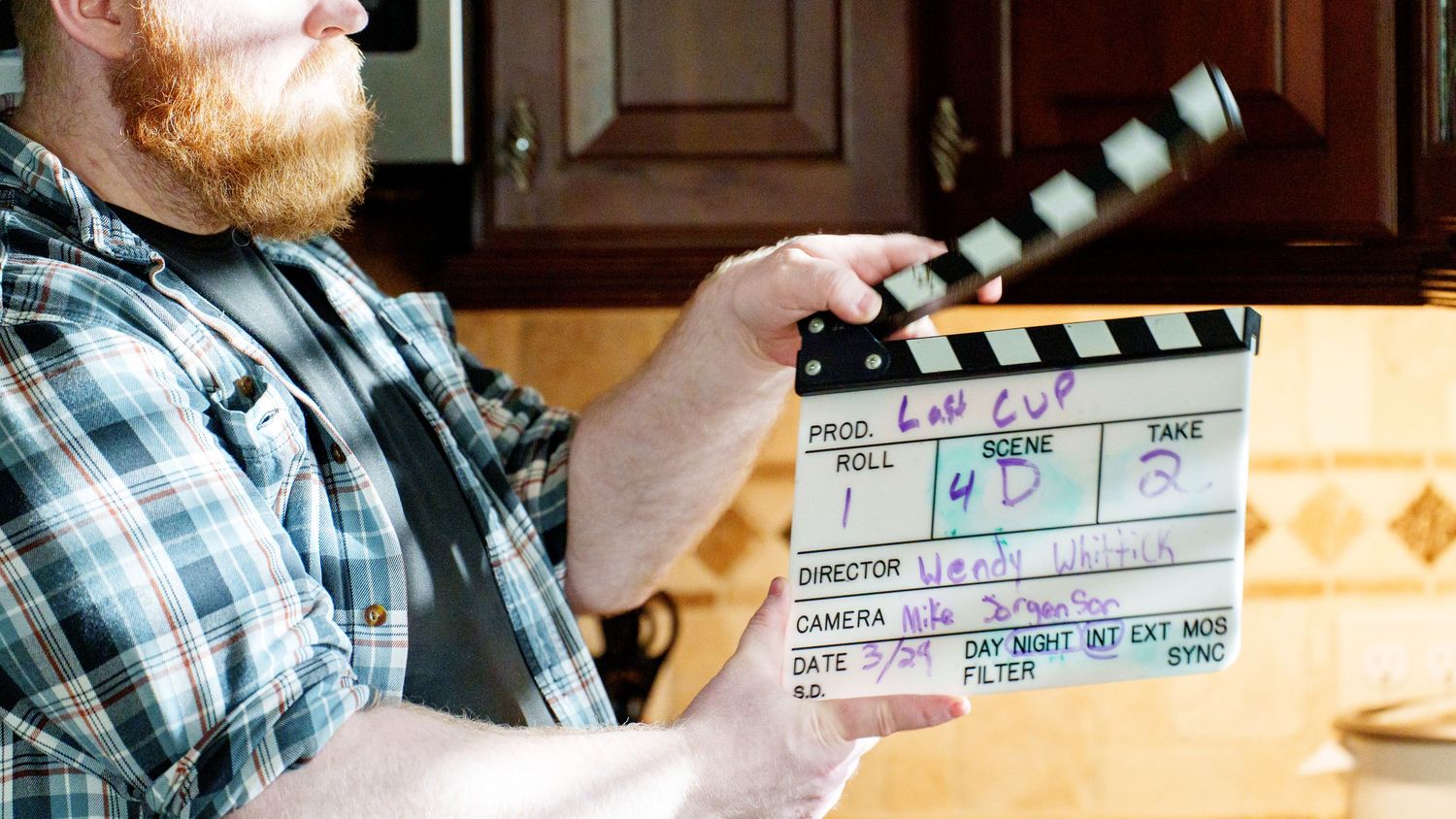 A bearded man holds a film clapperboard on a movie set in a kitchen, ready for a scene take.