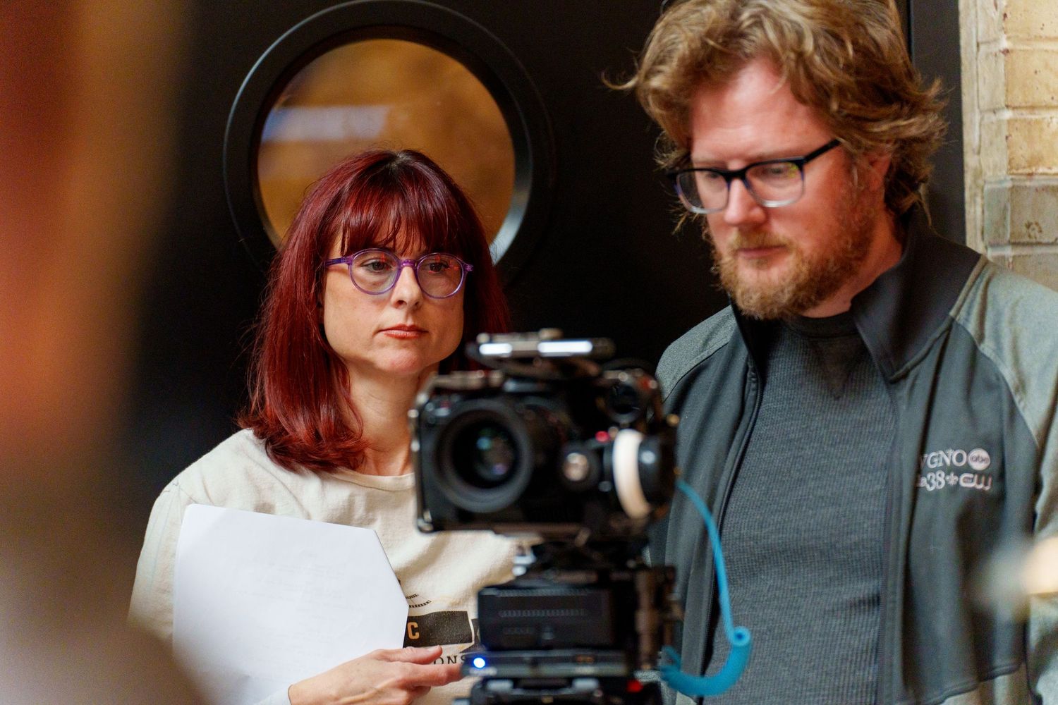 A woman with red hair and a man with a beard examine a professional camera on a film set.
