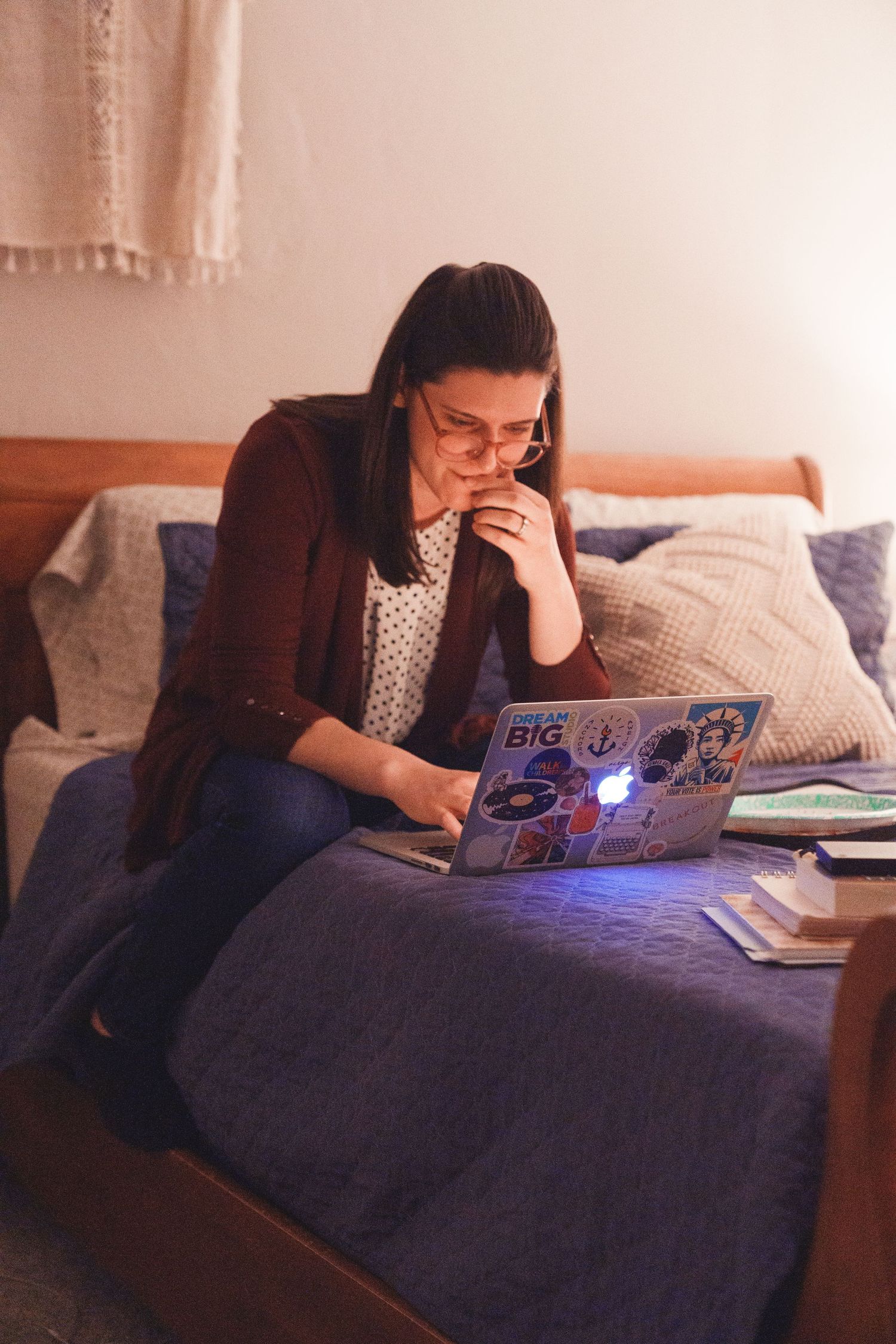 Woman in glasses sitting on bed working intently on a sticker-covered laptop in a warmly lit bedroom.