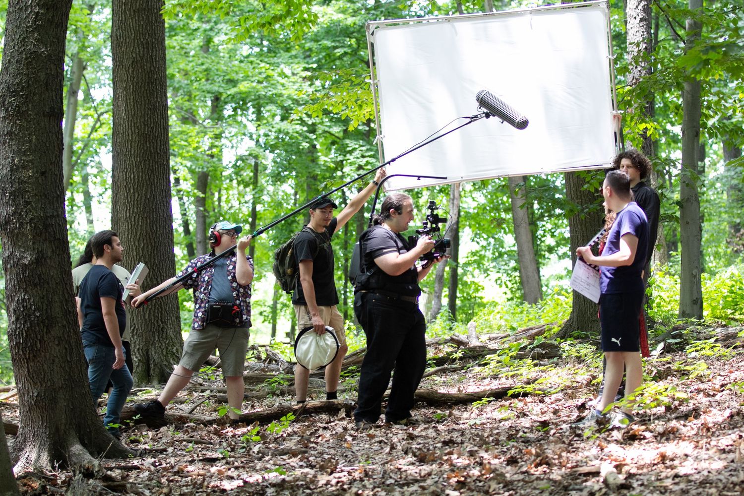 Film crew shooting outdoors in a forest with a boom mic and reflector board on a sunny day.