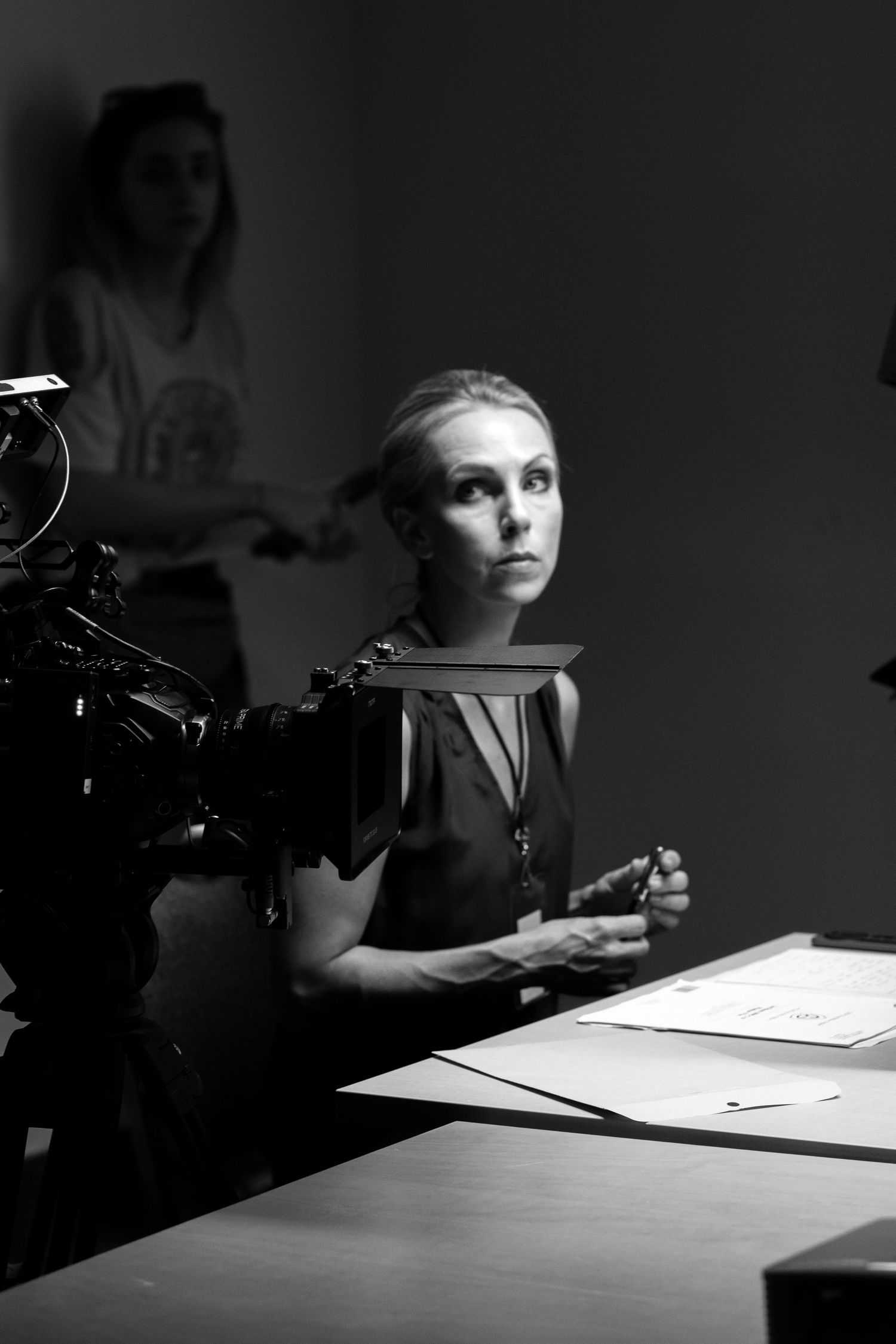 Black and white photo of a woman sitting at a desk on a film set with a camera nearby.