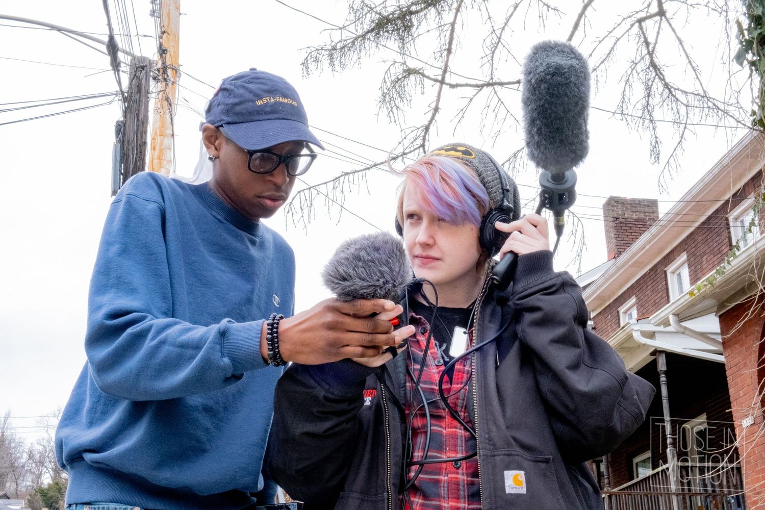 Two film students review audio equipment outdoors, one holding a boom mic while the other checks a recording device.