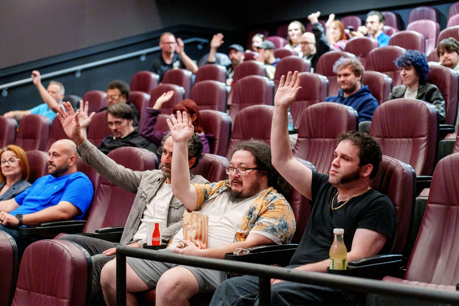 Movie theater audience members sitting in reddish seats watching and reacting with various expressions during a screening.