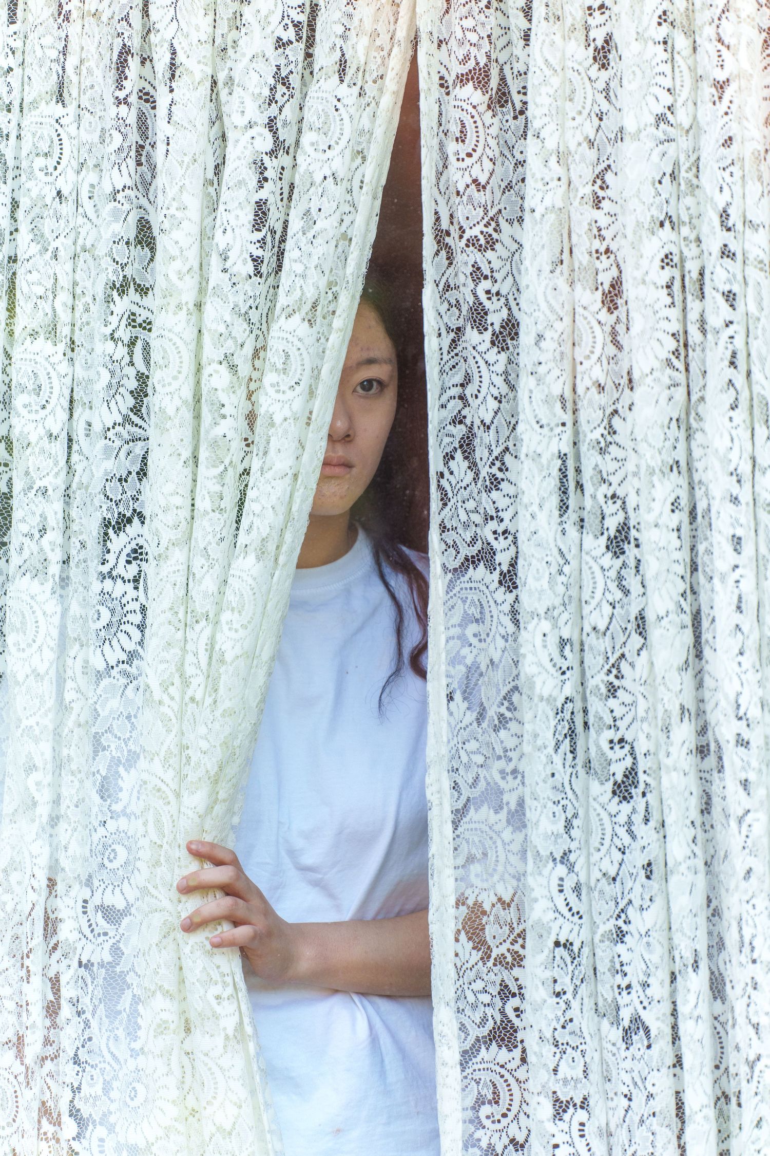 A figure partially visible through delicate white lace curtains in a bright setting.