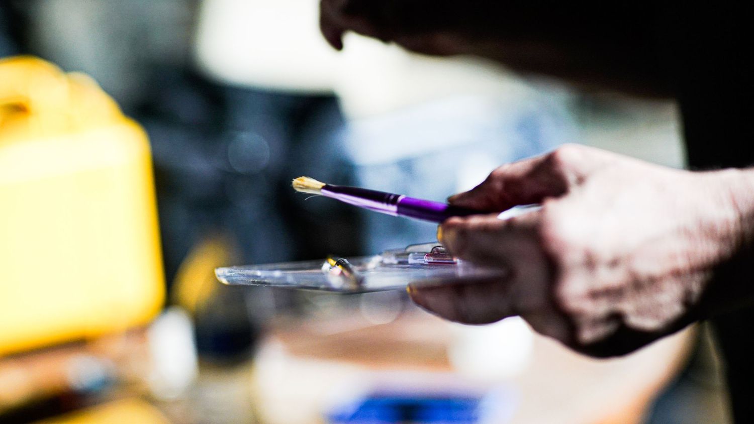 Close-up of hands holding a palette and purple-handled brush during a film production makeup session.