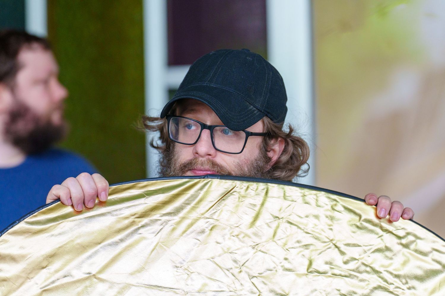 Man with glasses and black cap peeks over a gold reflector disc used in photography lighting outdoors.
