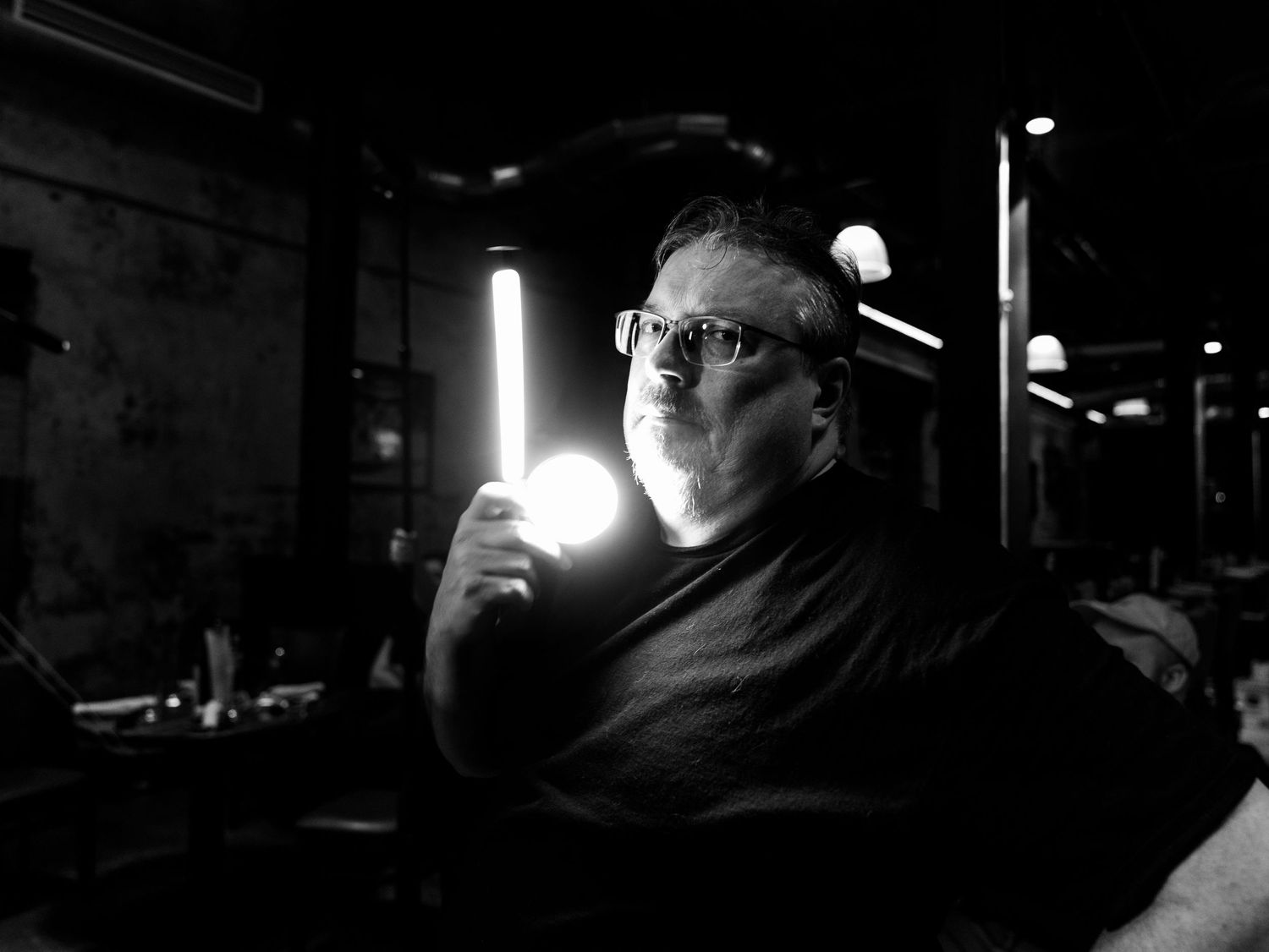 Black and white photo of a man holding a glowing light tube in a dark, moody studio setting.