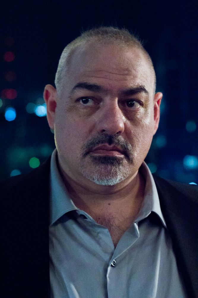 Serious middle-aged bald man with goatee in dark jacket against night city lights background.