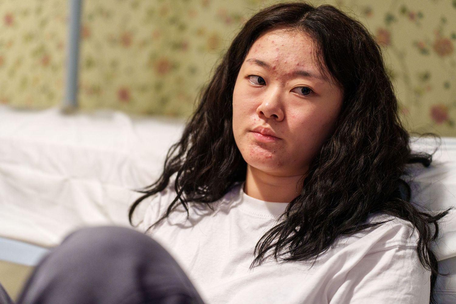 A young Asian woman with long wavy black hair sits in a hospital bed wearing a white patient gown, looking concerned.