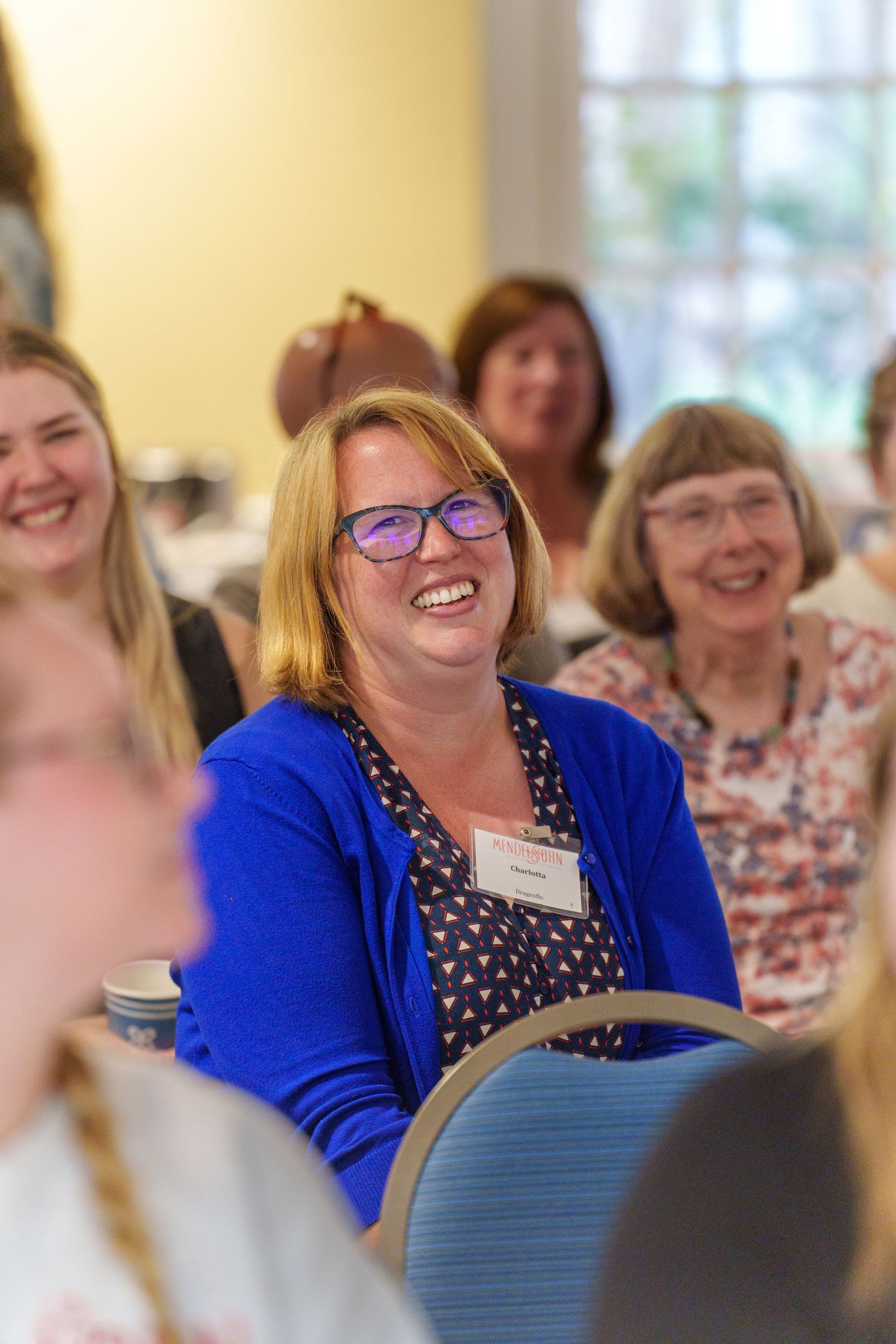 Women smiling and laughing at an indoor event, with a woman in a blue cardigan and glasses in focus.
