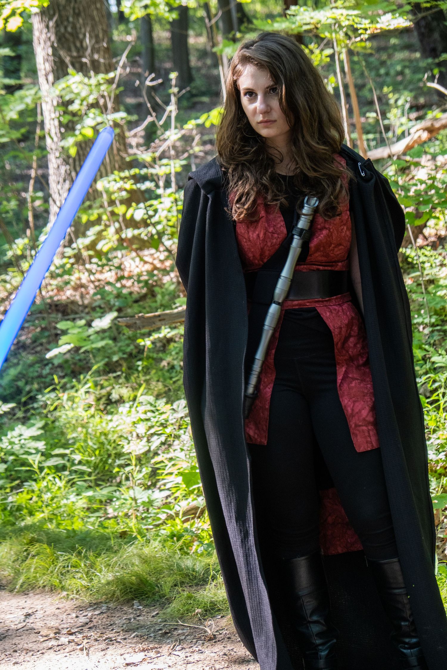 A cosplayer in dark robes and red accents posing with a lightsaber in a wooded area.