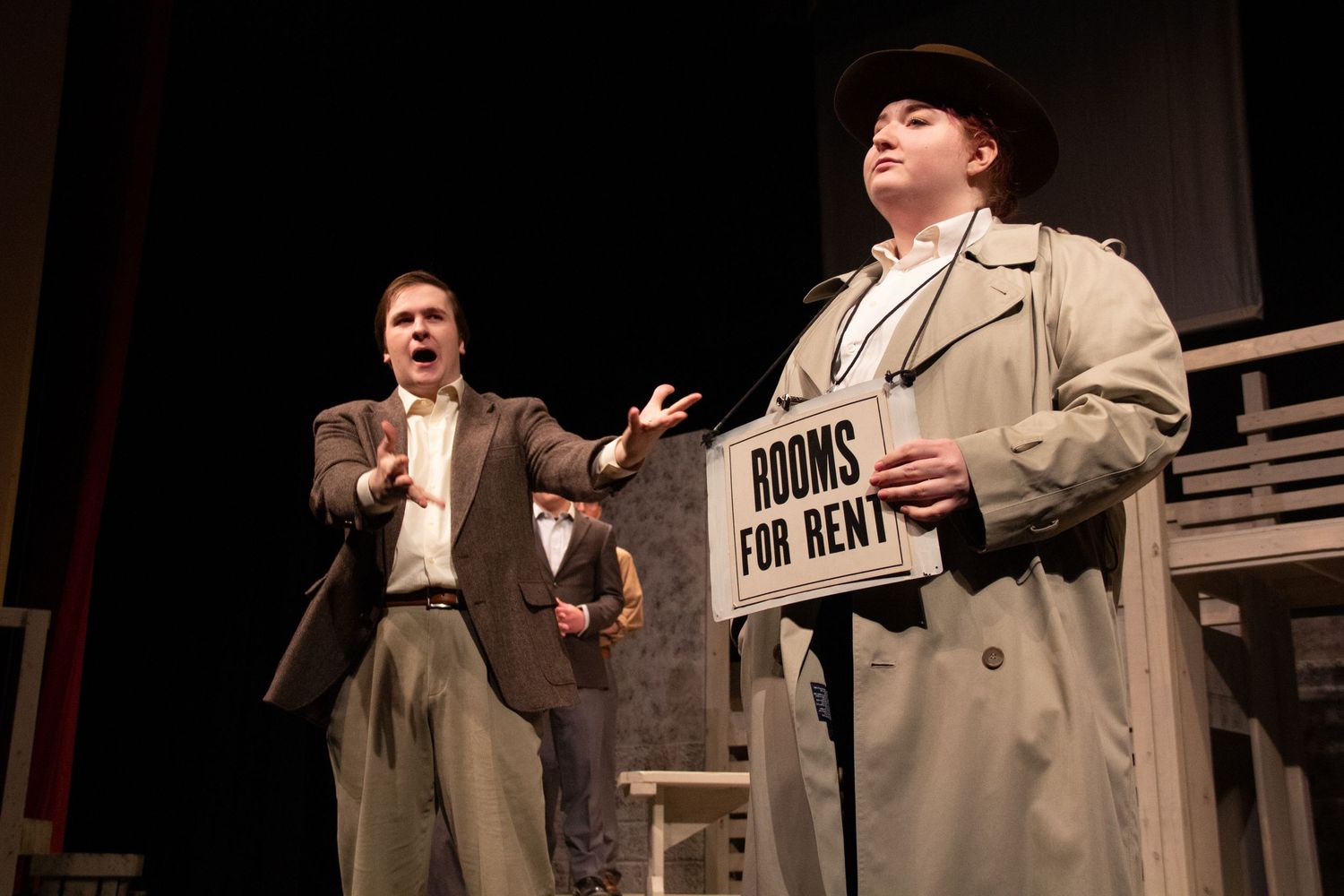 Stage actors performing in trench coats with 'For Rent' sign visible in background.