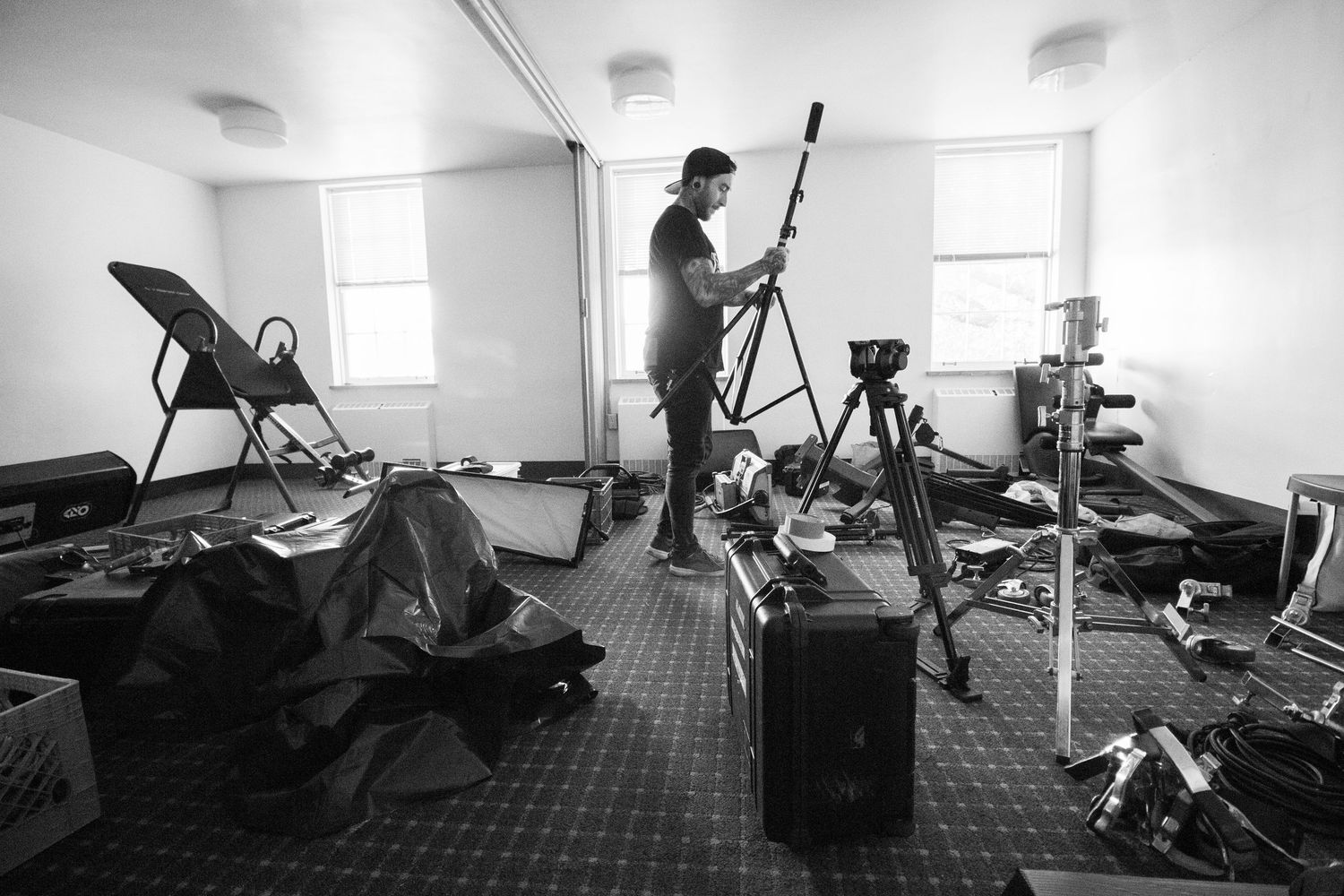 Black and white photo of a man setting up film equipment in a cluttered room with tripods and speakers.