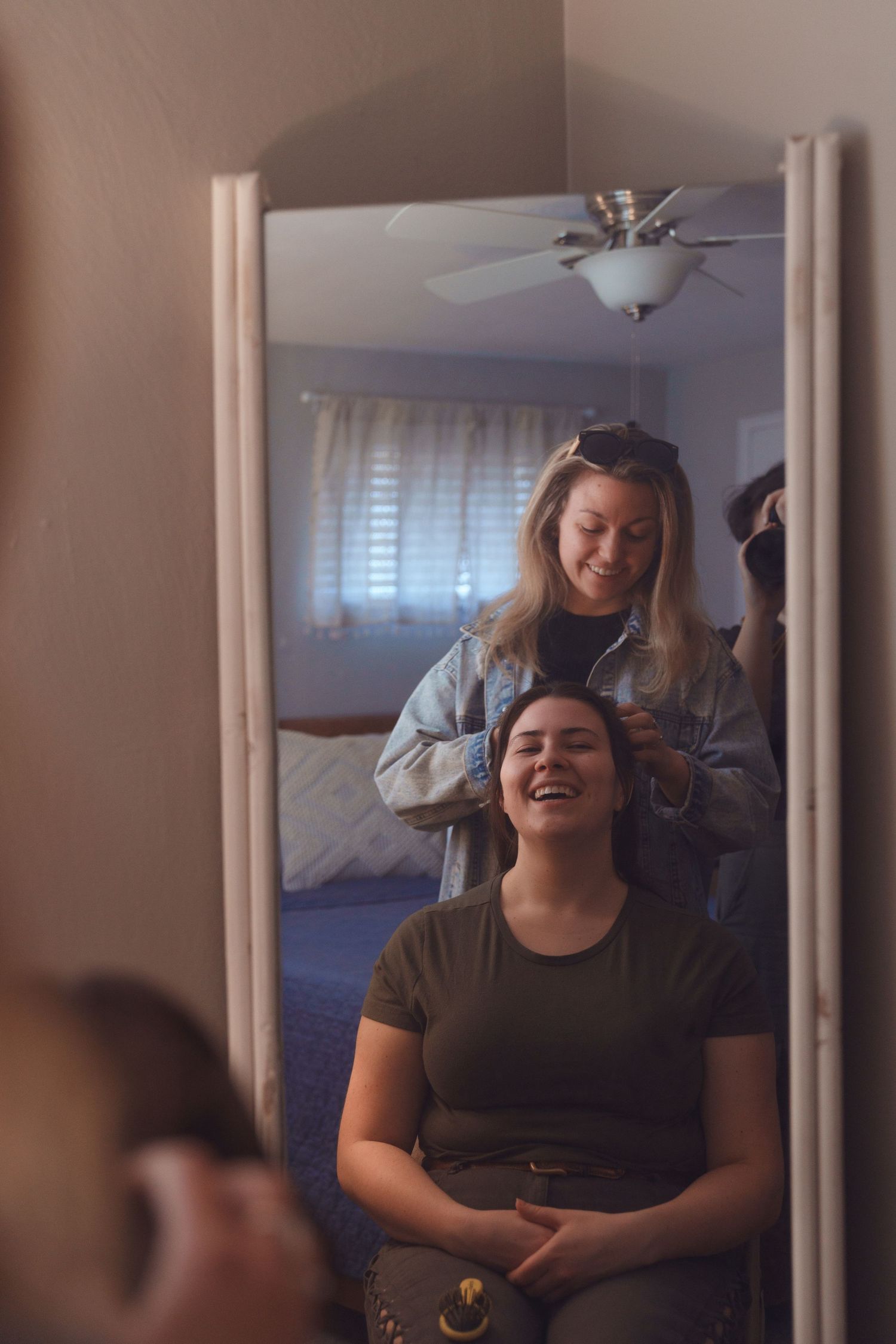 Woman laughs joyfully as friend styles her hair in bedroom mirror reflection.
