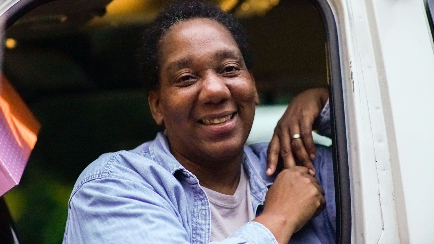 A smiling Black woman in a light blue shirt leans out of a vehicle window outdoors.