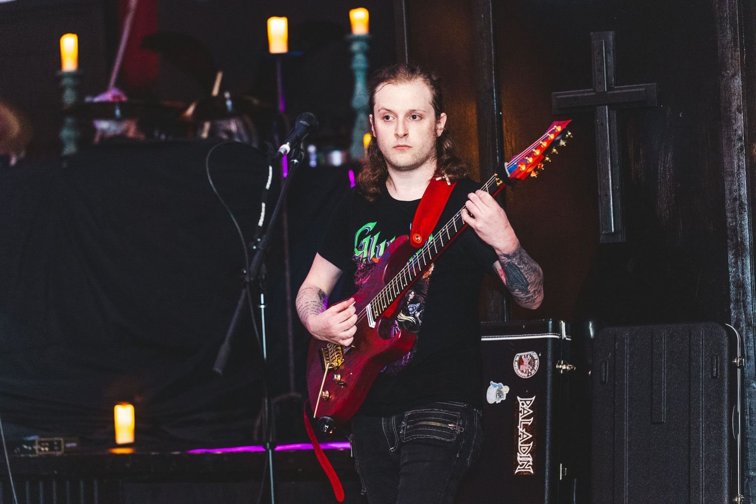 A musician performs on stage with a red electric guitar under dim lighting with glowing yellow lamps in the background.