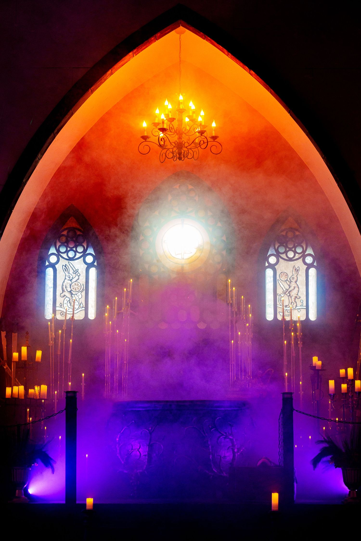 Colorful illuminated church interior with arched ceiling glowing in orange, purple and blue lights through stained glass windows.