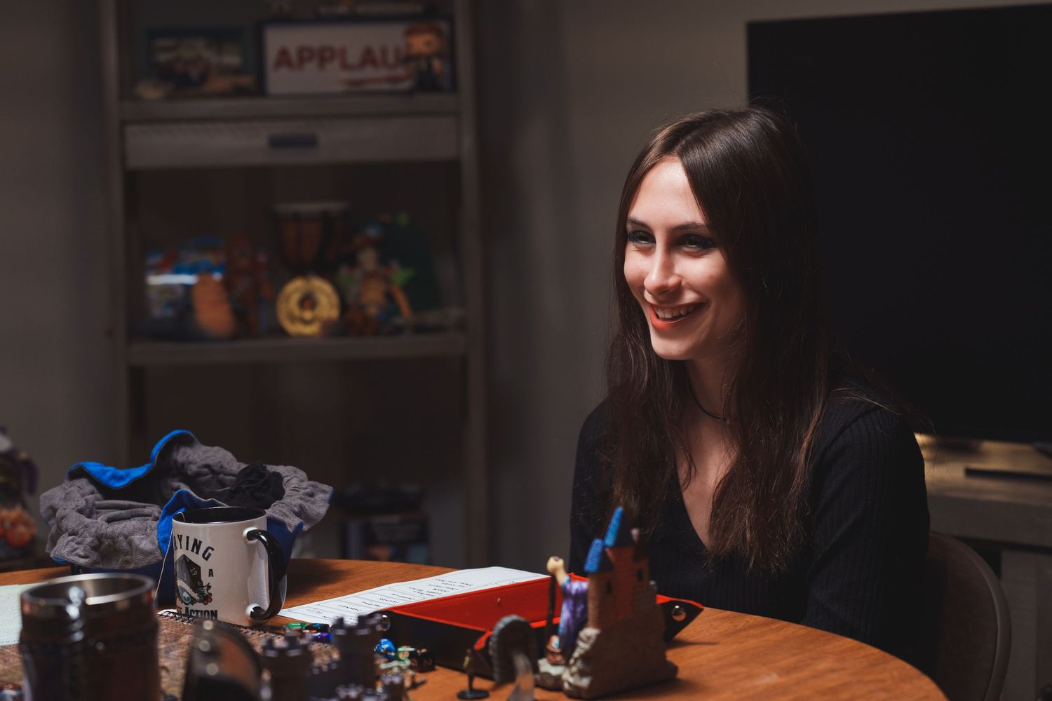 Smiling woman with long dark hair sitting at a table with gaming miniatures and dice in a dimly lit room.