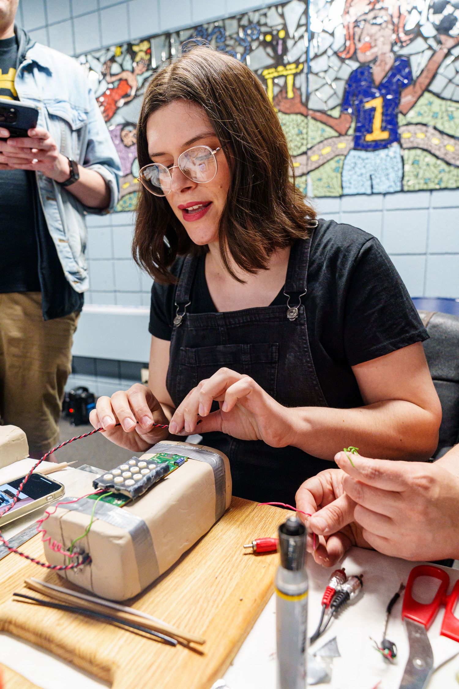 Woman with glasses works on electronics project at a table with circuit boards and wiring components.
