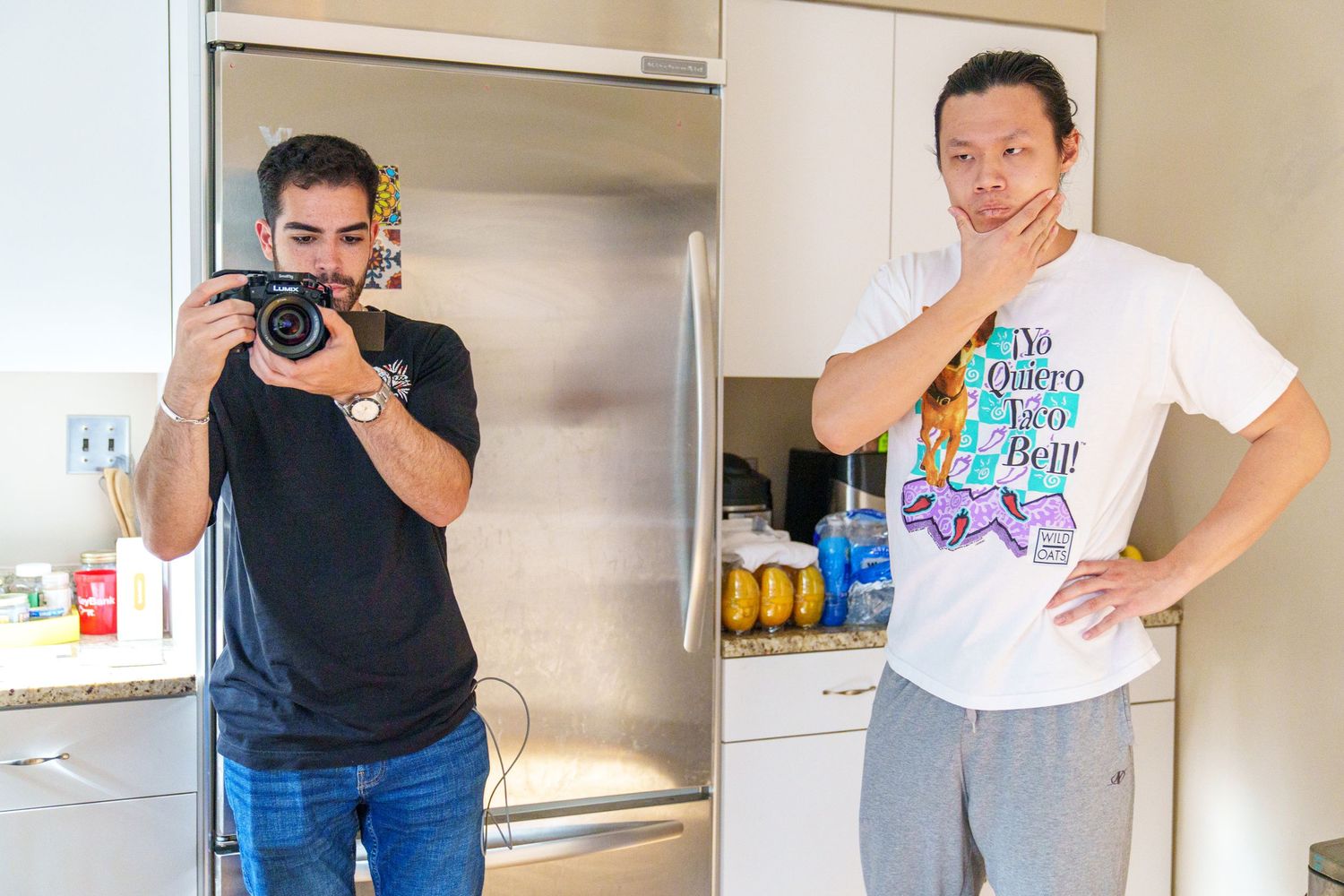 Two men in a kitchen, one photographing with a DSLR camera, another in a graphic tee posing thoughtfully.