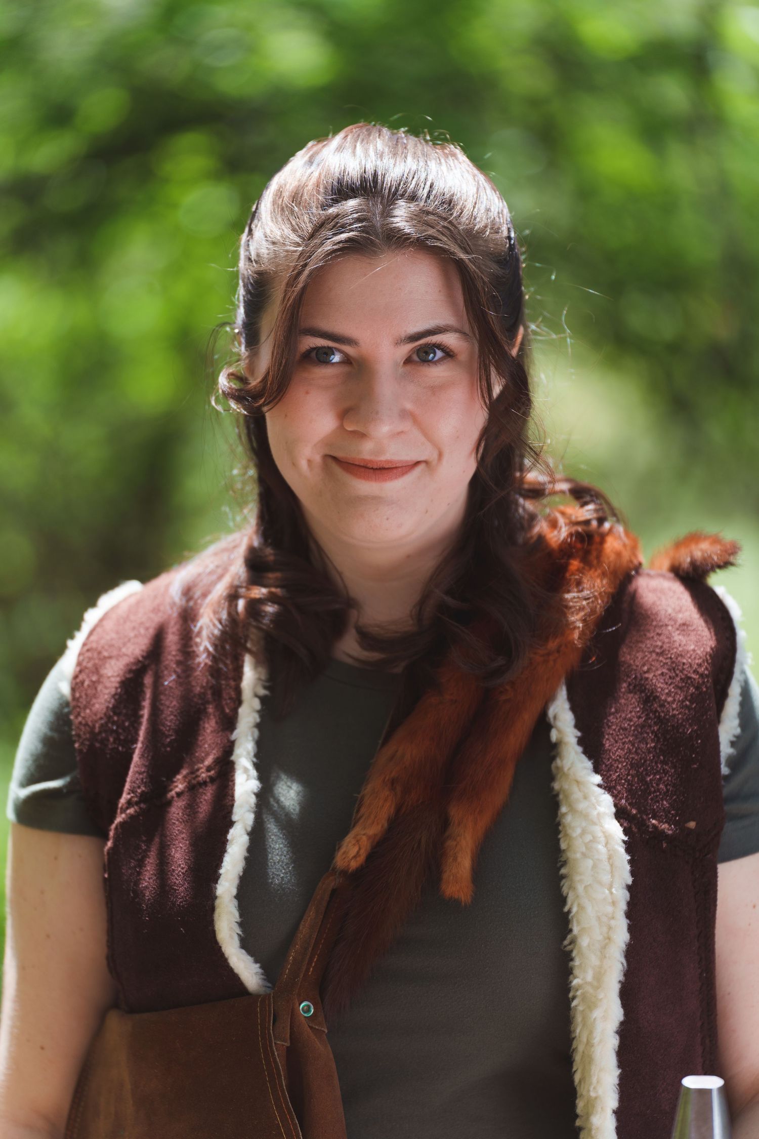 Young woman smiling outdoors, wearing a brown sherpa vest with a squirrel on her shoulder, green trees in background.