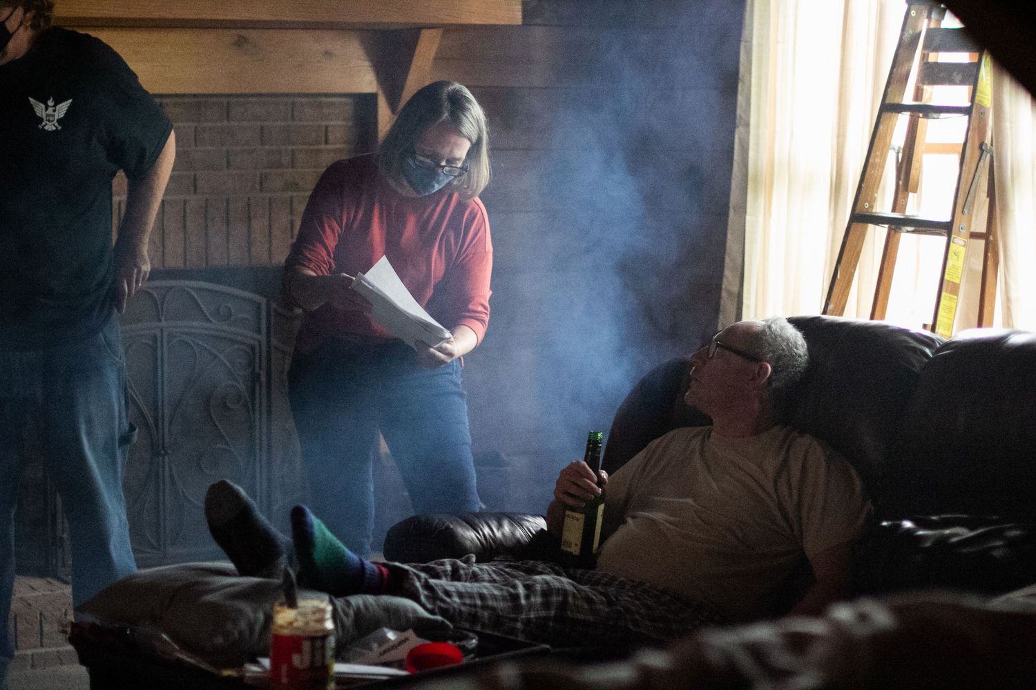 Two people in a smoky rustic cabin, one lying on a couch while another stands reading papers near a fireplace.