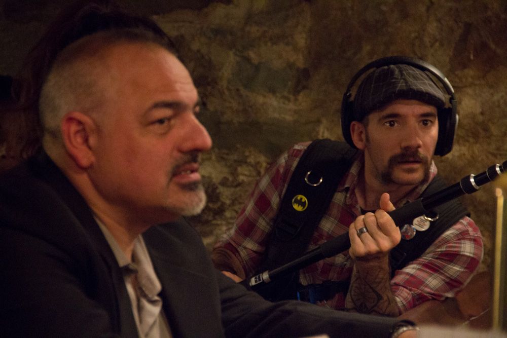 Two men with mustaches sit together in a dimly lit room during a discussion.