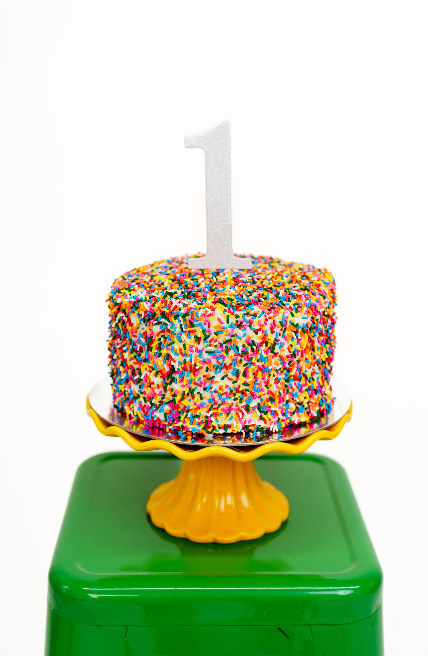 Birthday cake covered in rainbow sprinkles with white number one topper on green and yellow stand.