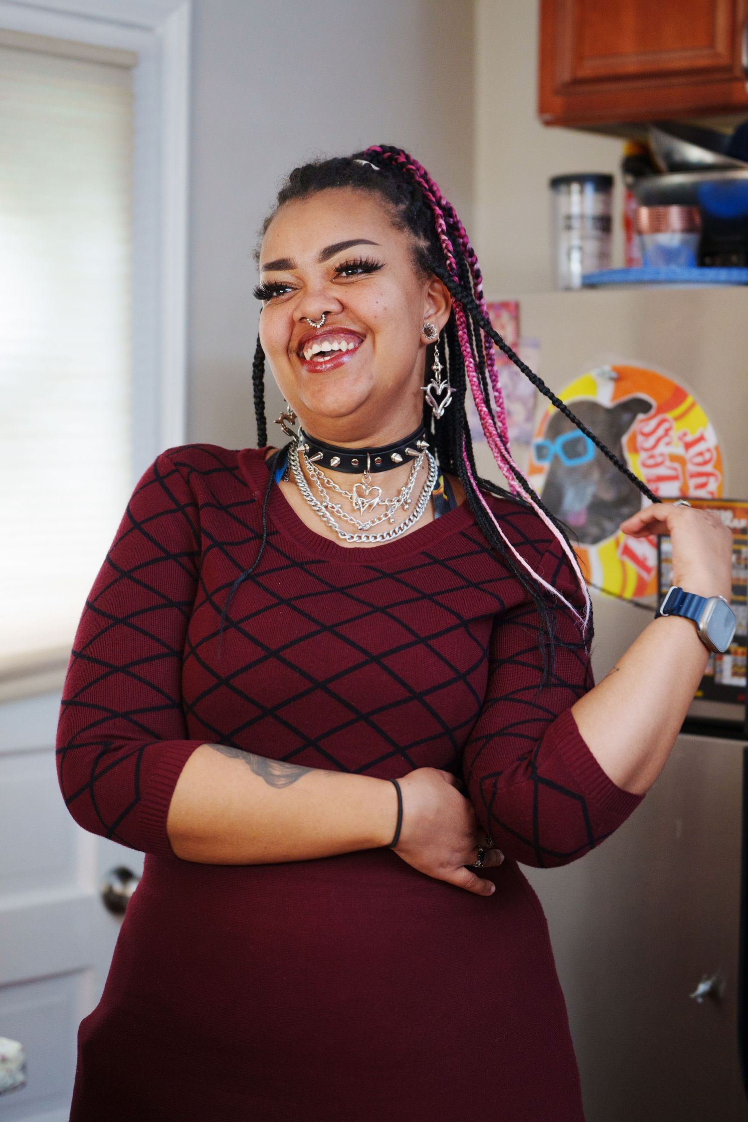 Smiling woman with pink braids and layered necklaces wearing a maroon dress, posing confidently in a kitchen.
