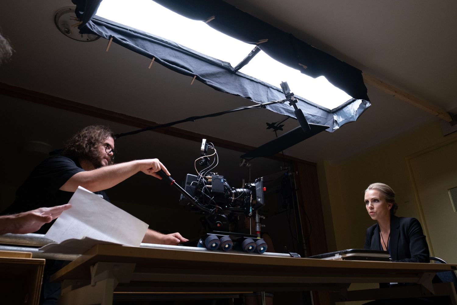 A filmmaker reviews script pages while a camera is aimed at an actress on a film set.