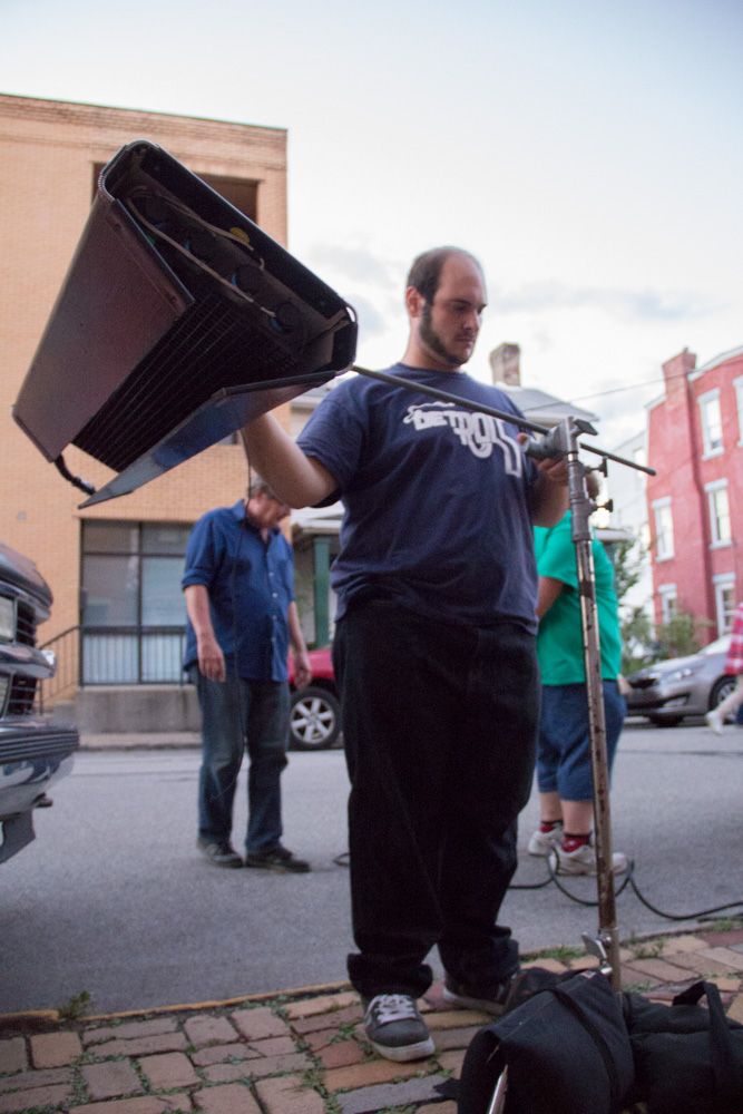 Man in Detroit t-shirt holds film lighting equipment on a cobblestone street during an outdoor video production shoot.