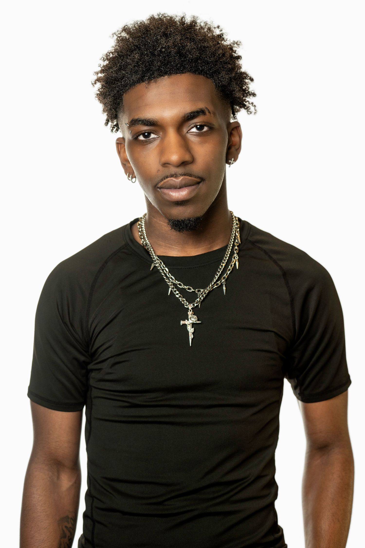 Young Black man in black t-shirt wearing gold cross necklace and chain, posing against white background.