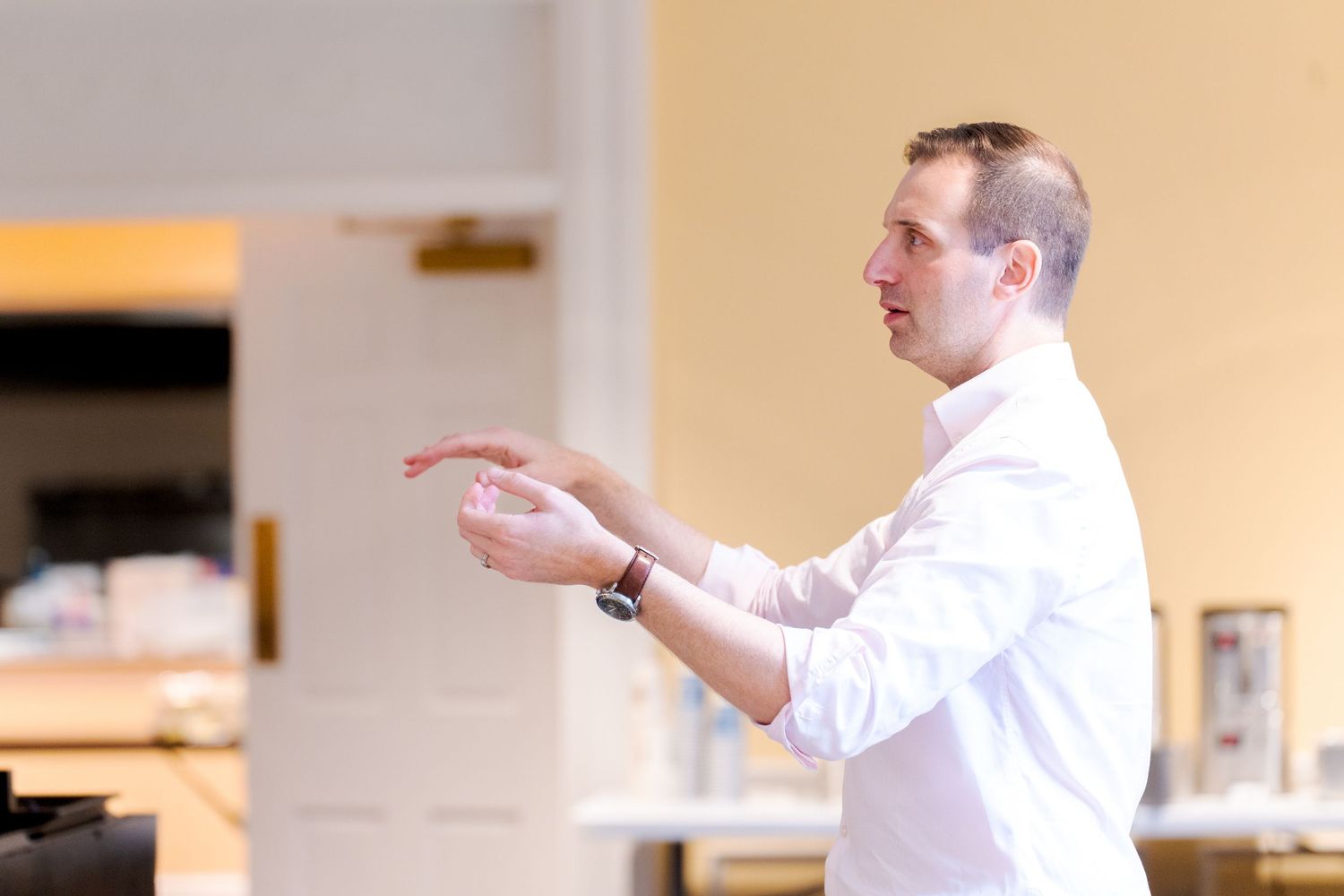 A person in a white shirt gestures while speaking to a group in an indoor space.