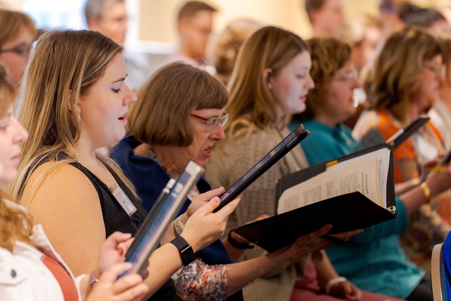 Women sing from sheet music binders during a choir performance in a sunlit church setting.