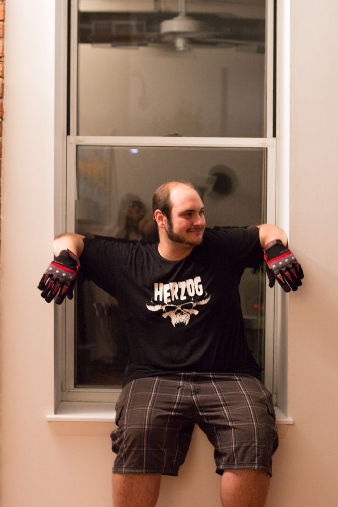 Man in Herzog t-shirt and motorcycle gloves posing by a window in plaid shorts, smiling indoors.