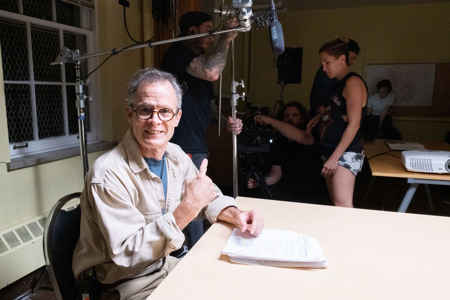 An elderly man smiles and gives a thumbs up while sitting at a desk on a film set with crew members in the background.