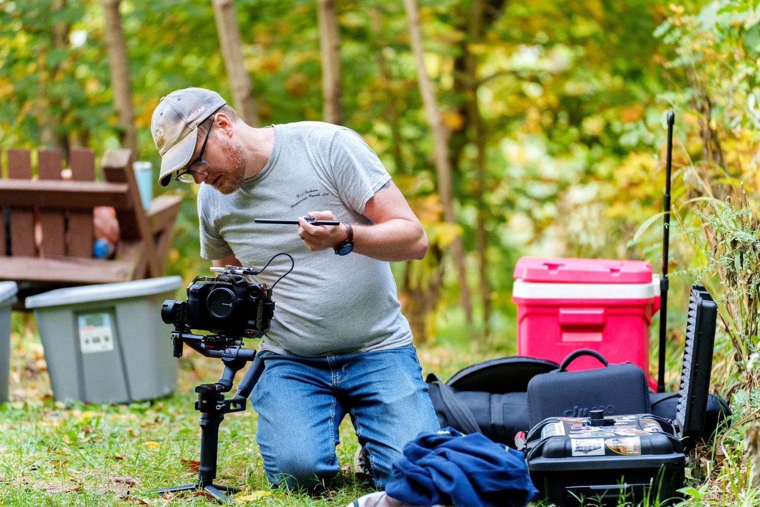 Videographer adjusting camera on gimbal stabilizer outdoors with equipment cases and red cooler in background.
