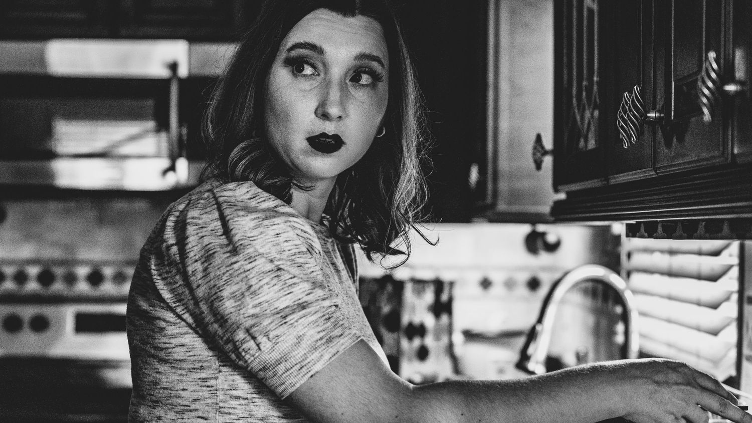 A black and white image shows a person in a floral dress looking upward while standing in a vintage-style kitchen.