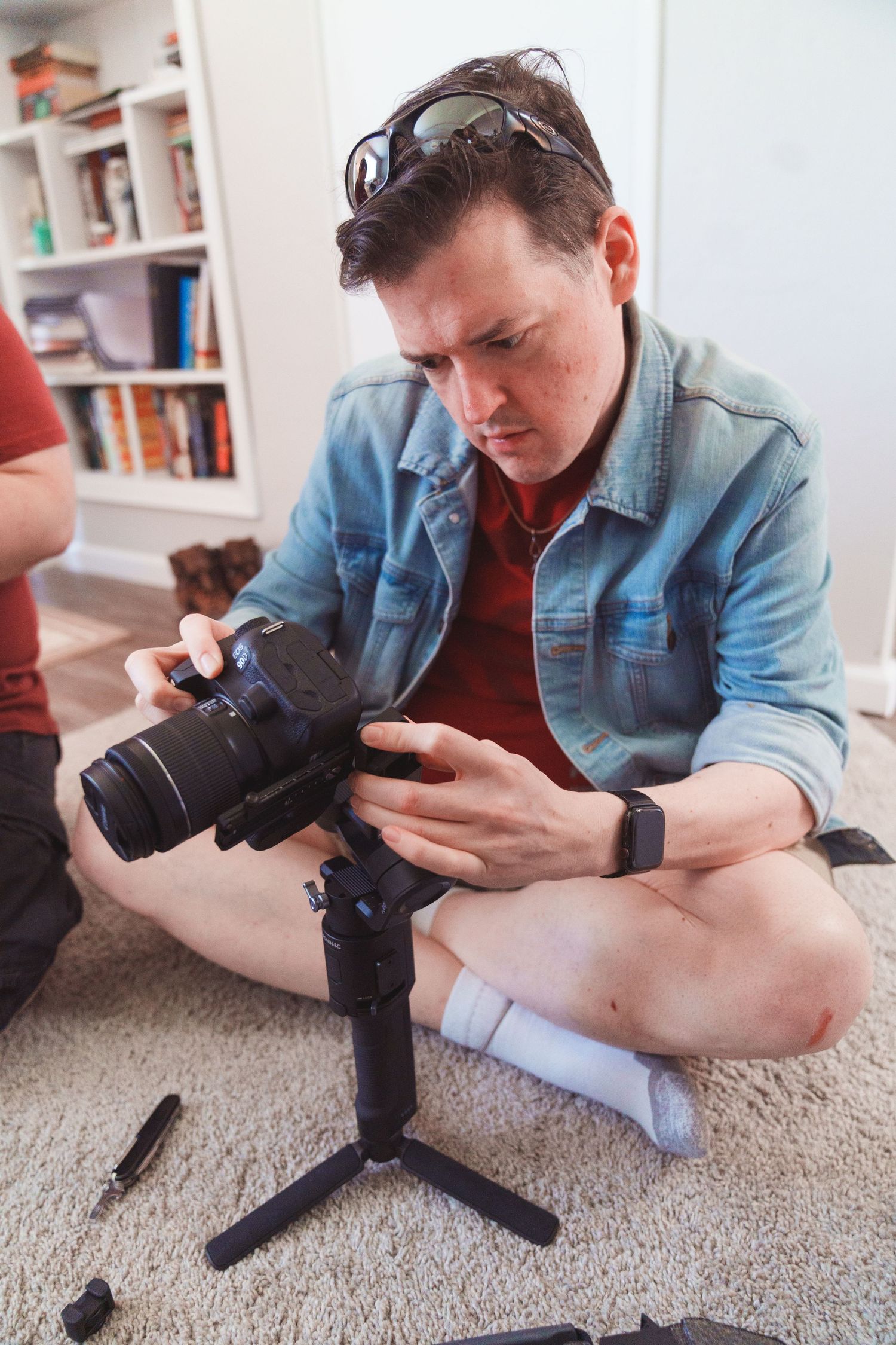 Person in denim jacket examining camera equipment while sitting on carpeted floor.