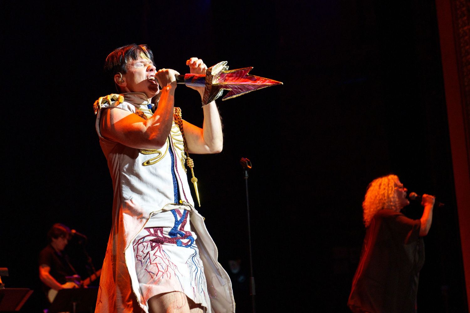 Performers on stage during a theatrical show with dramatic lighting and microphones.