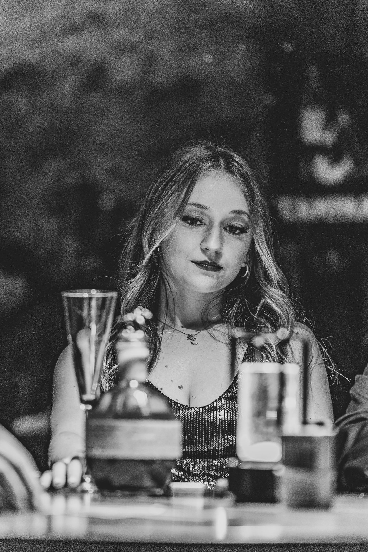 A black and white portrait of someone in a sparkly top sitting at a restaurant table with drinking glasses.
