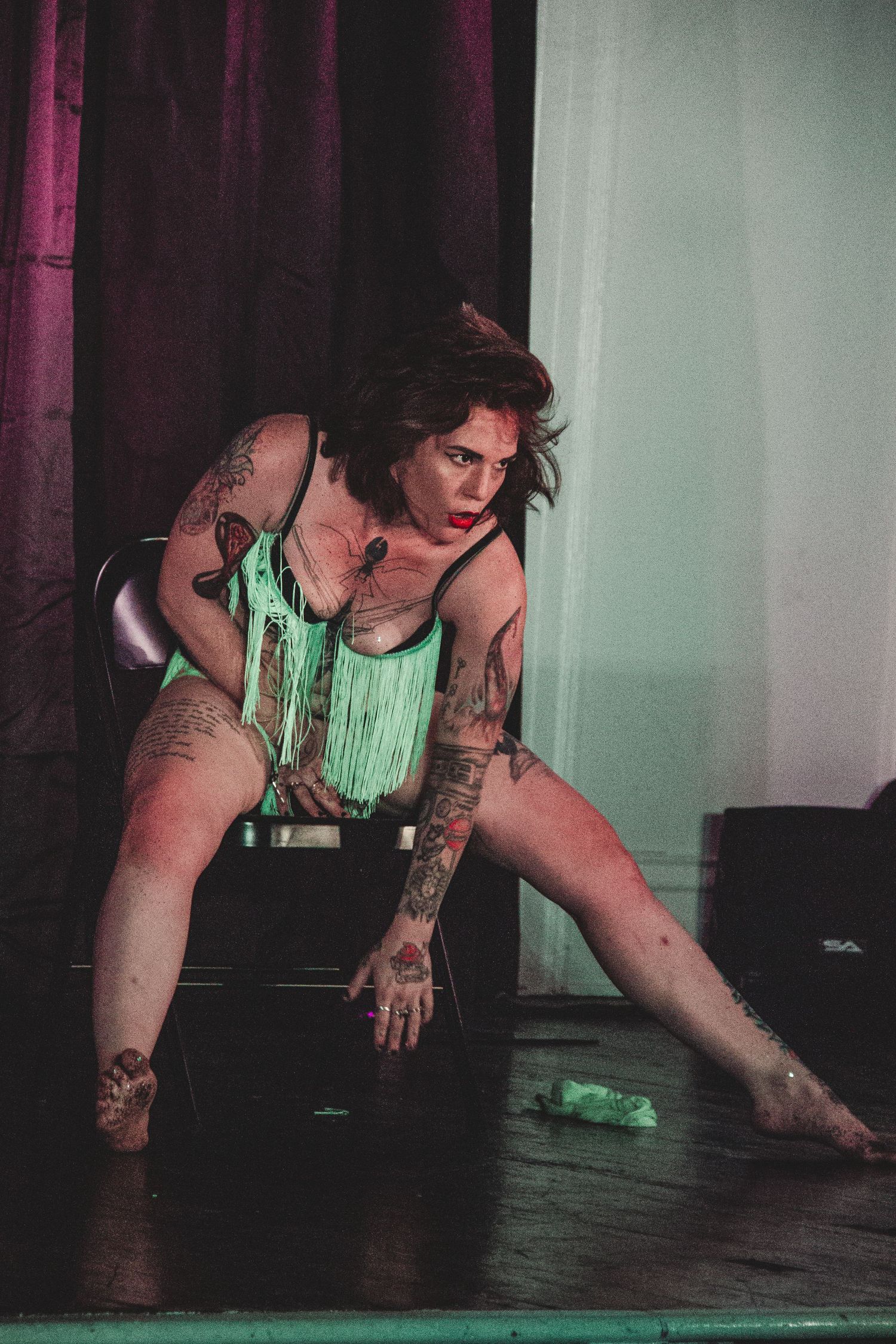 Tattooed performer in green fringe costume crouches on dark stage.