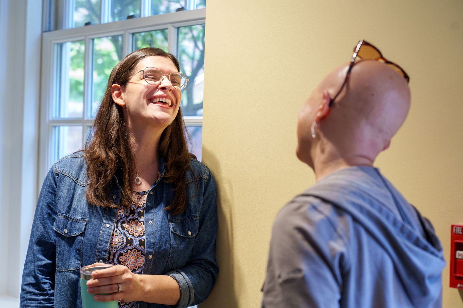 Two people laughing and conversing indoors near a window, one wearing glasses and a denim jacket.