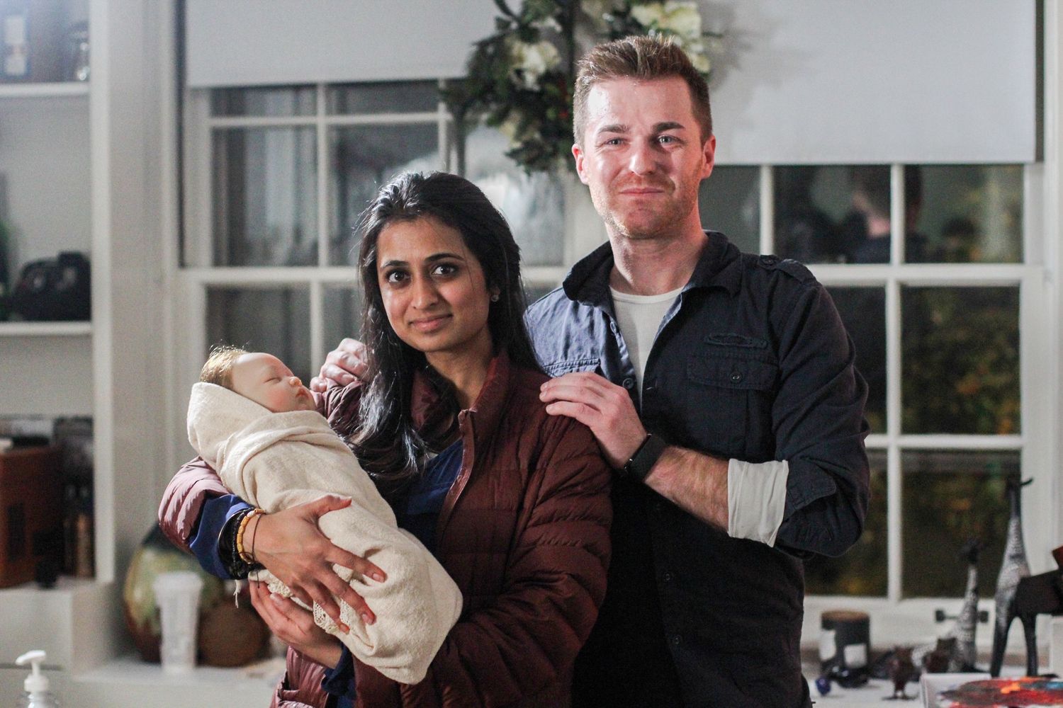 A couple smiling and holding a newborn baby indoors, posing together in a warmly lit room.