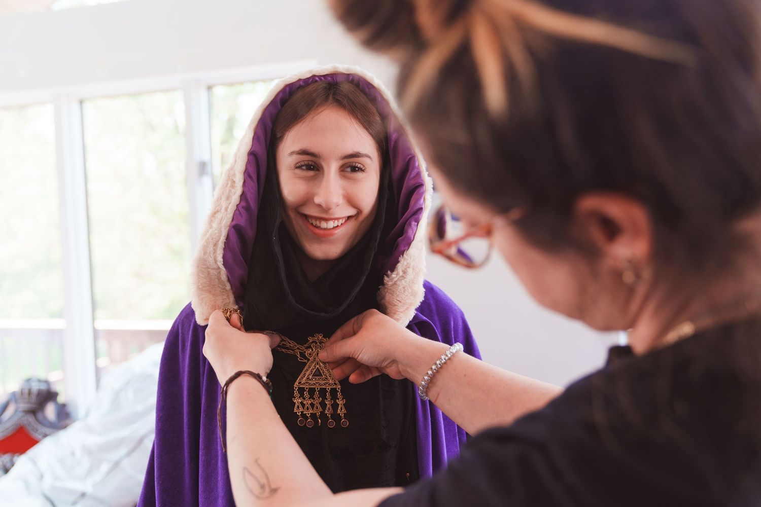 Woman smiling as another person adjusts a purple hijab and gold necklace on her.