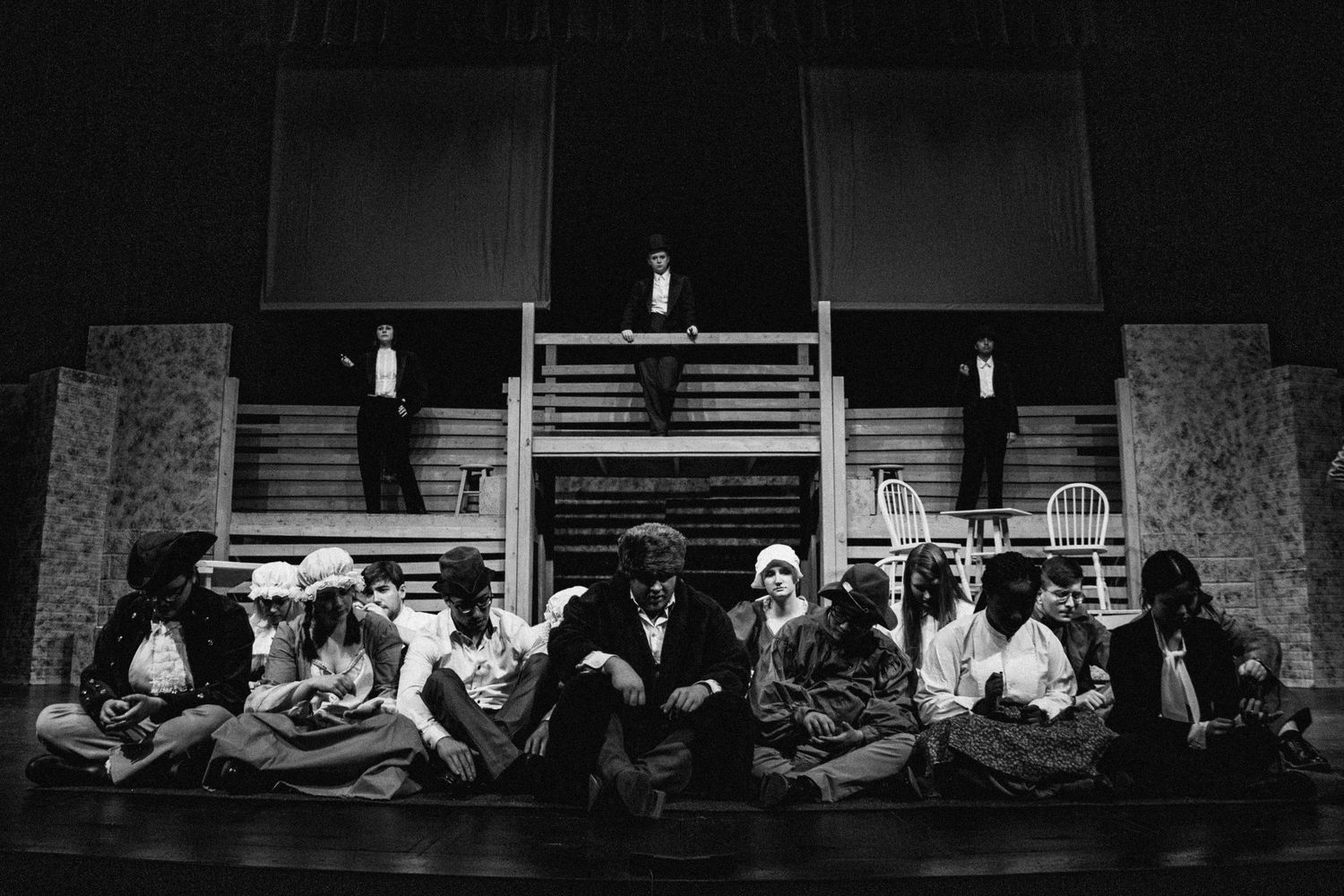 Black and white theatrical scene with performers arranged across dark stage.