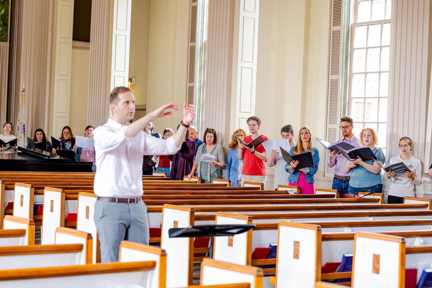 A choir director conducts singers in a bright church sanctuary during a choral rehearsal.
