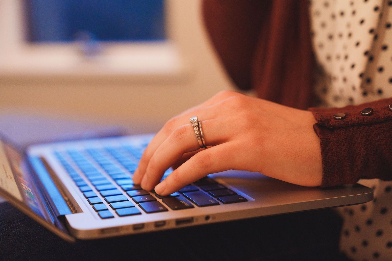 Close-up of a woman's hand wearing a ring while typing on a MacBook laptop keyboard.