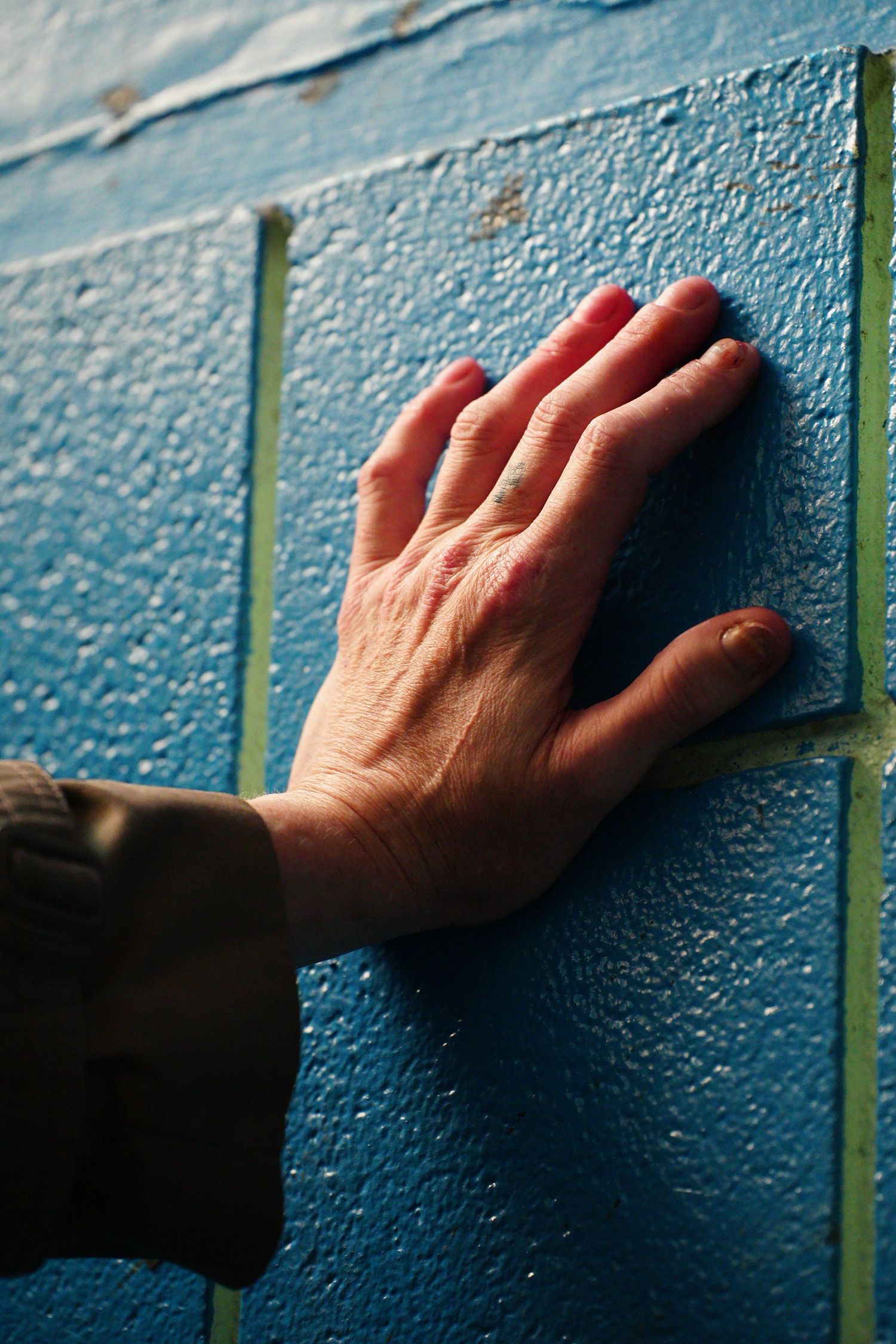 A weathered hand pressed against a textured blue painted brick wall, wearing a dark sleeve.