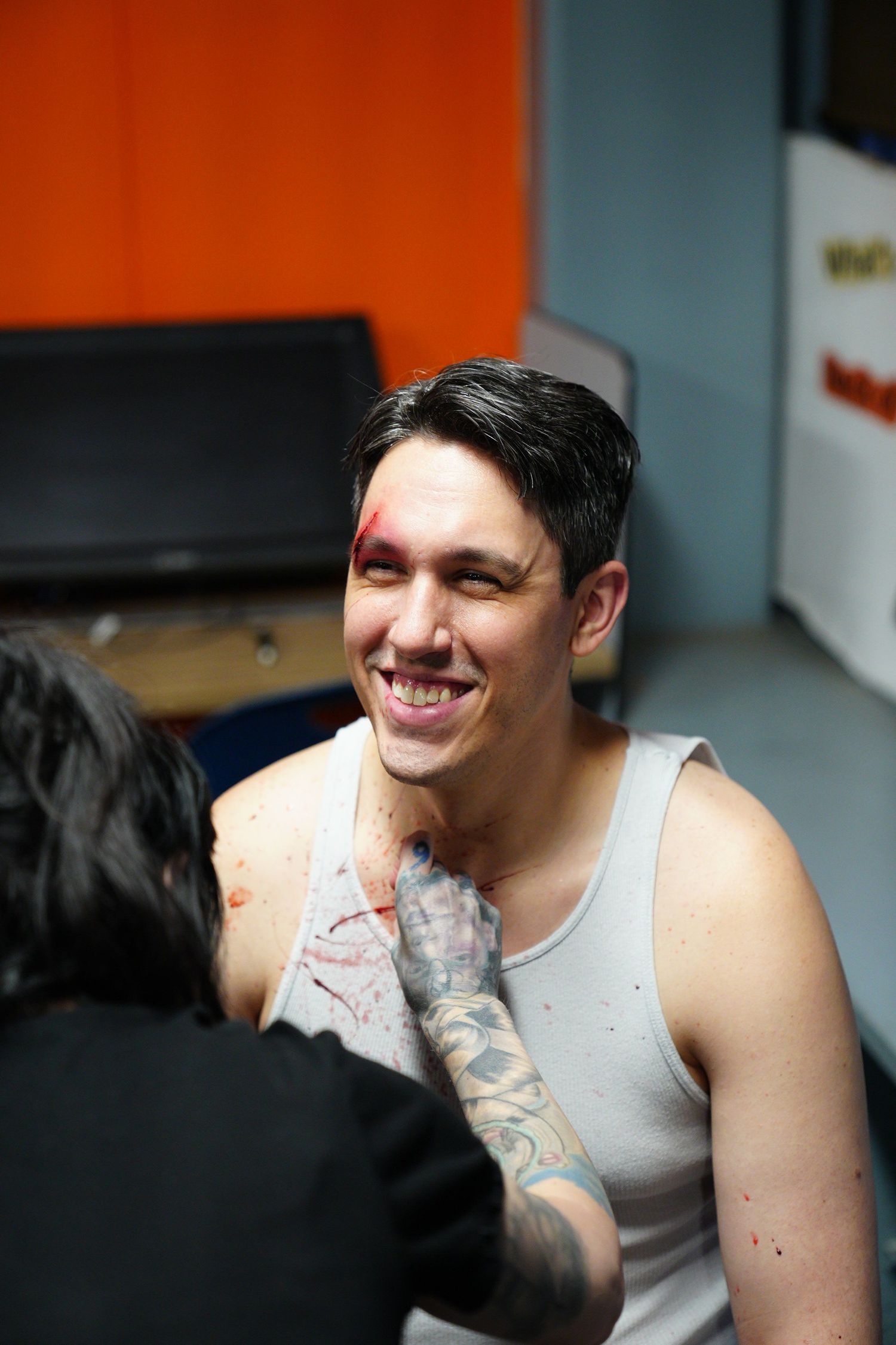 Smiling tattooed man in tank top with fake blood makeup being applied by makeup artist indoors.